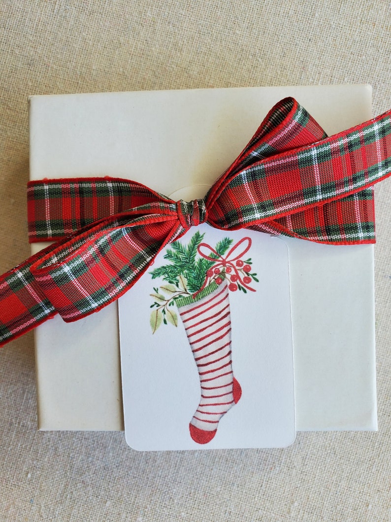 May include: A white gift box with a red and green plaid ribbon tied in a bow. A white gift tag with a red and white striped stocking with green holly and red berries is attached to the ribbon.