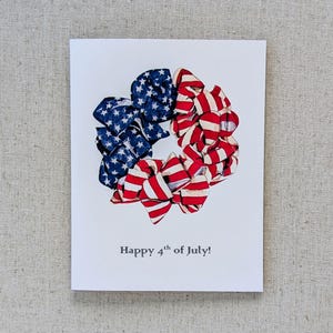 May include: A white card with a red, white, and blue fabric wreath in the center. The wreath is made of fabric strips with stars and stripes. The text "Happy 4th of July!" is printed below the wreath.