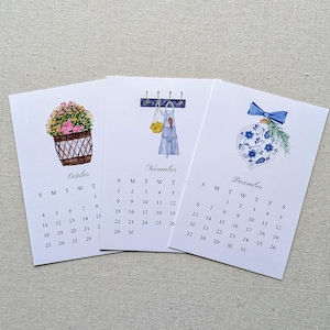 May include: Three watercolor calendars for October, November, and December. Each calendar features a different illustration: a flower basket, a kitchen scene, and a Christmas ornament. The calendars display the days of the month.