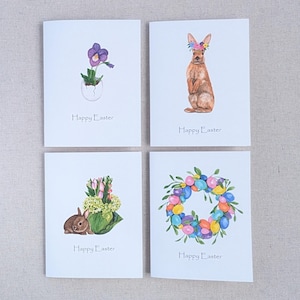 May include: Set of four Easter greeting cards with watercolor illustrations. The cards feature a purple flower in a cracked egg, a brown rabbit wearing a flower crown, a brown rabbit with a bouquet of tulips and a wreath of colorful eggs.