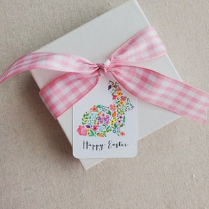 Puede incluir: Una caja de regalo blanca con una cinta de cuadros rosa atada en un lazo. Una etiqueta de regalo con un dise&ntilde;o de conejo floral y el texto "Feliz Pascua" est&aacute; unida a la caja.