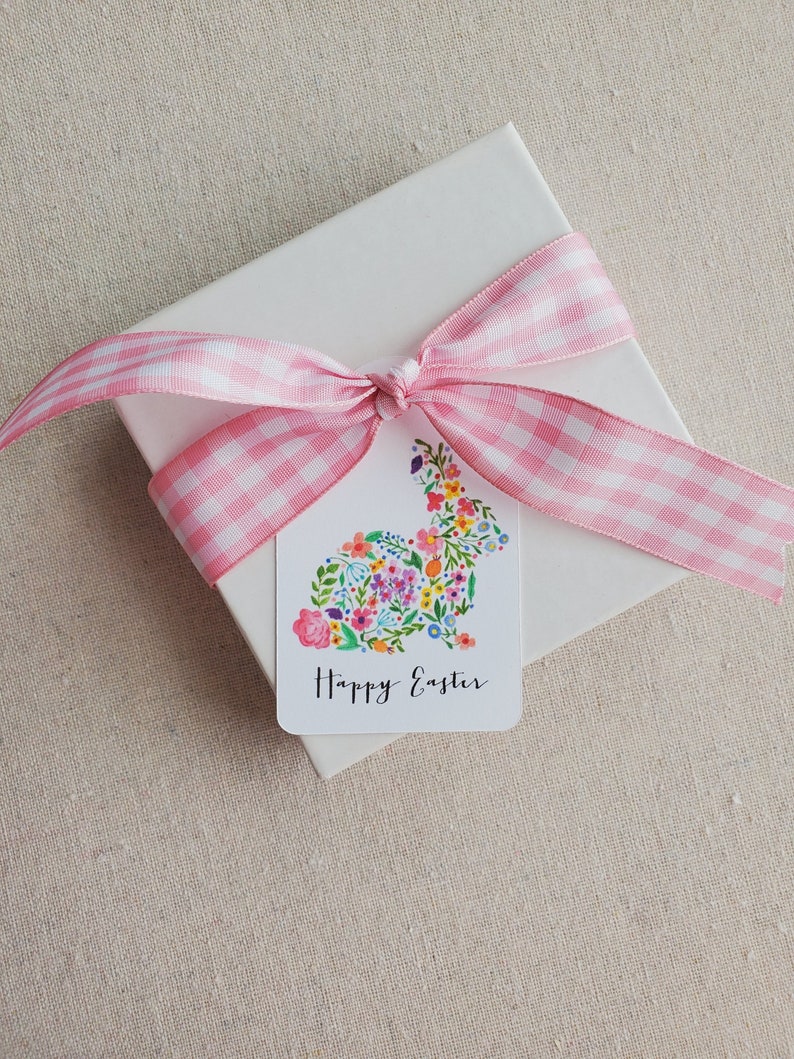 Puede incluir: Una caja de regalo blanca con una cinta de cuadros rosa atada en un lazo. Una etiqueta de regalo con un dise&ntilde;o de conejo floral y el texto "Feliz Pascua" est&aacute; unida a la caja.