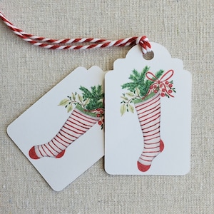 May include: Two white gift tags with red and white striped stockings, green holly, and red berries. The tags are tied together with a red and white twine.