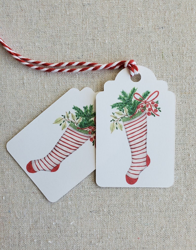 May include: Two white gift tags with red and white striped stockings, green holly, and red berries. The tags are tied together with a red and white twine.