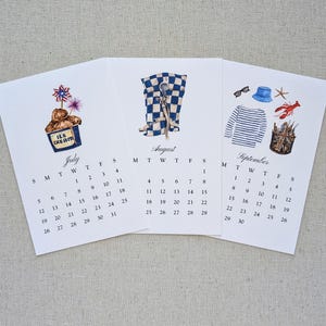 May include: Three seasonal calendar pages with watercolor illustrations. July features ice cream, August shows a checkered pattern with scissors, and September displays nautical elements. Each page includes a month name and calendar dates.