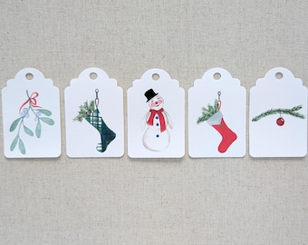 Könnte beinhalten: Fünf weiße Geschenkanhänger mit verschiedenen Weihnachtsmotiven. Die Anhänger zeigen einen Schneemann, einen Strumpf, Mistelzweig, einen Weihnachtsbaumzweig mit rotem Ornament und einen Strumpf mit grünem Karomuster.