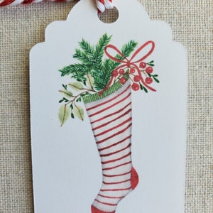 May include: A white gift tag with a red and white striped stocking filled with green holly and red berries. The stocking has a red bow and is tied with a red and white twine.