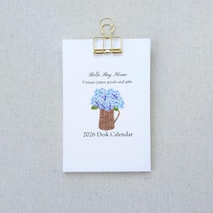 May include: A white desk calendar clipped with a gold binder clip. The calendar features the text "Bella Bay Home" and "Unique paper goods and gifts." A watercolor illustration of blue hydrangeas in a woven basket is below the text, with "2026 Desk Calendar" at the bottom.