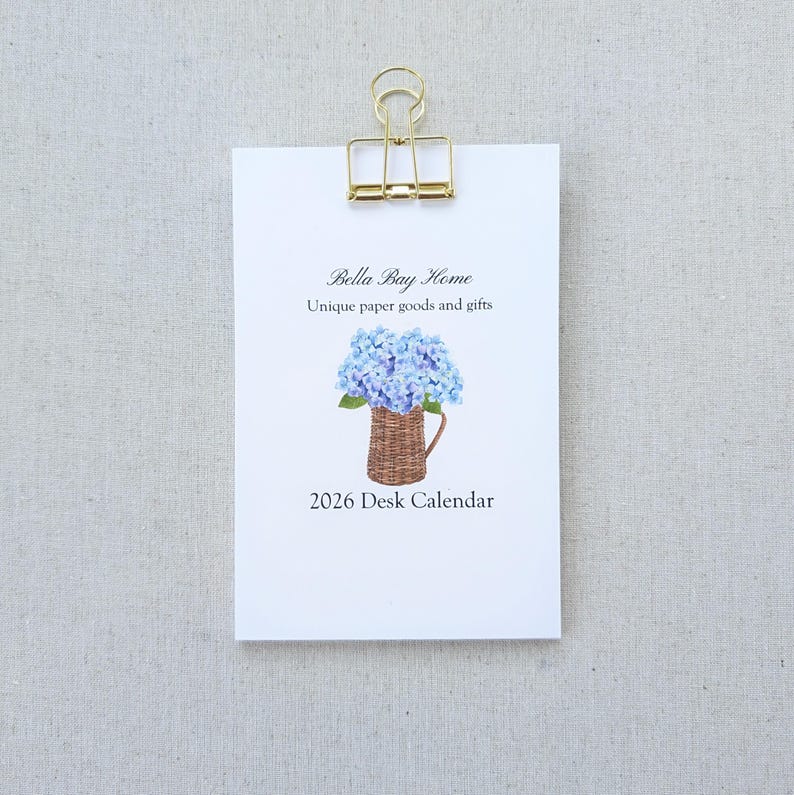 May include: A white desk calendar clipped with a gold binder clip. The calendar features the text "Bella Bay Home" and "Unique paper goods and gifts." A watercolor illustration of blue hydrangeas in a woven basket is below the text, with "2026 Desk Calendar" at the bottom.