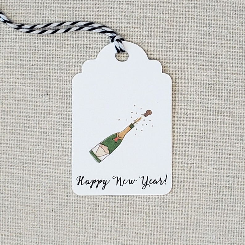 May include: White gift tag with a black and white striped string. The tag features a hand-drawn illustration of a champagne bottle with a cork popping out and the text "Happy New Year!"