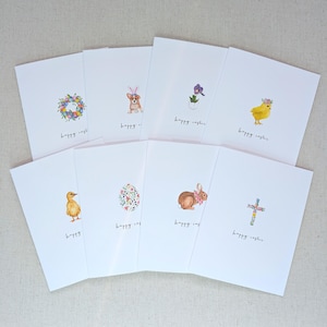 May include: A collection of white Easter greeting cards with watercolor illustrations. Designs include a wreath, a corgi with bunny ears, a flower, a chick, a duckling, a decorated egg, a bunny, and a cross. Each card has the text "happy easter".