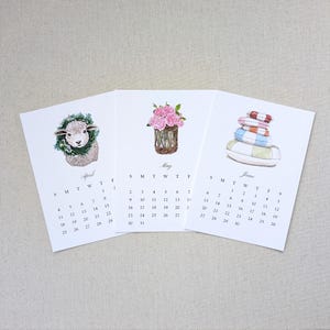 May include: Three white calendar pages for April, May, and June. April features a lamb with a green wreath, May shows pink flowers in a woven basket, and June displays a stack of colorful striped cushions. Each page includes a monthly grid.