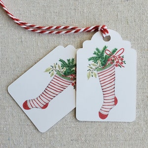 May include: Two white gift tags with red and white striped stockings filled with green holly and red berries. The tags are tied together with red and white twine.