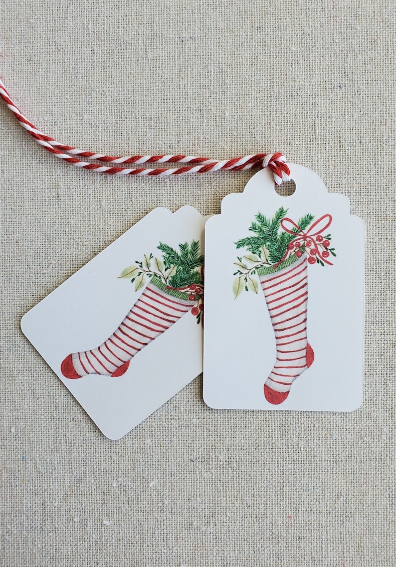 May include: Two white gift tags with red and white striped stockings filled with green holly and red berries. The tags are tied together with red and white twine.