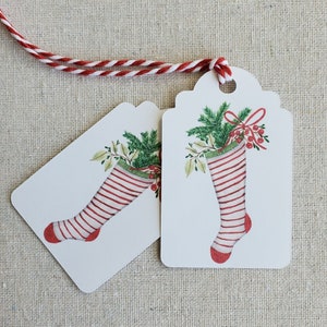 May include: Two white gift tags with scalloped edges and red and white striped stockings with green holly and red berries. The tags are tied together with a red and white twine.