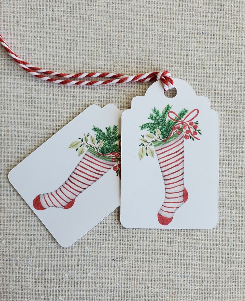 May include: Two white gift tags with scalloped edges and red and white striped stockings with green holly and red berries. The tags are tied together with a red and white twine.