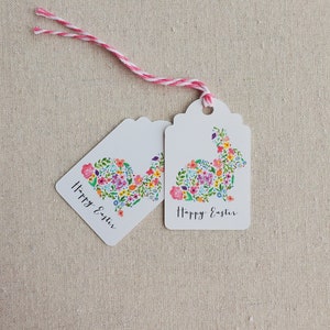 Puede incluir: Dos etiquetas de regalo blancas con un dise&ntilde;o de conejo floral y el texto "Feliz Pascua" en cada una. Las etiquetas est&aacute;n atadas juntas con un cordel rosa y blanco.