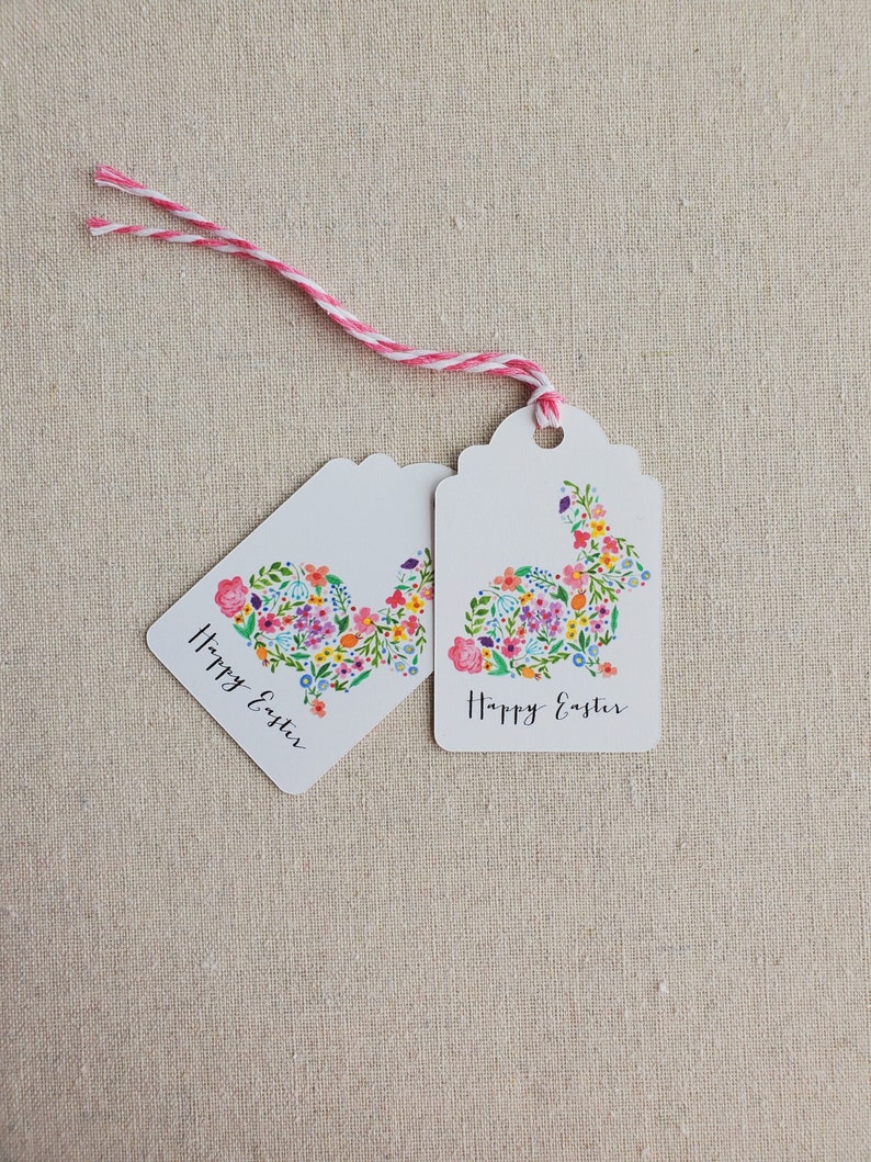 Puede incluir: Dos etiquetas de regalo blancas con un dise&ntilde;o de conejo floral y el texto "Feliz Pascua" en cada una. Las etiquetas est&aacute;n atadas juntas con un cordel rosa y blanco.