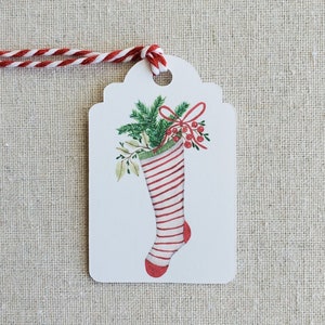 May include: A white gift tag with a red and white striped stocking filled with green holly and red berries. The tag is tied with a red and white twine.