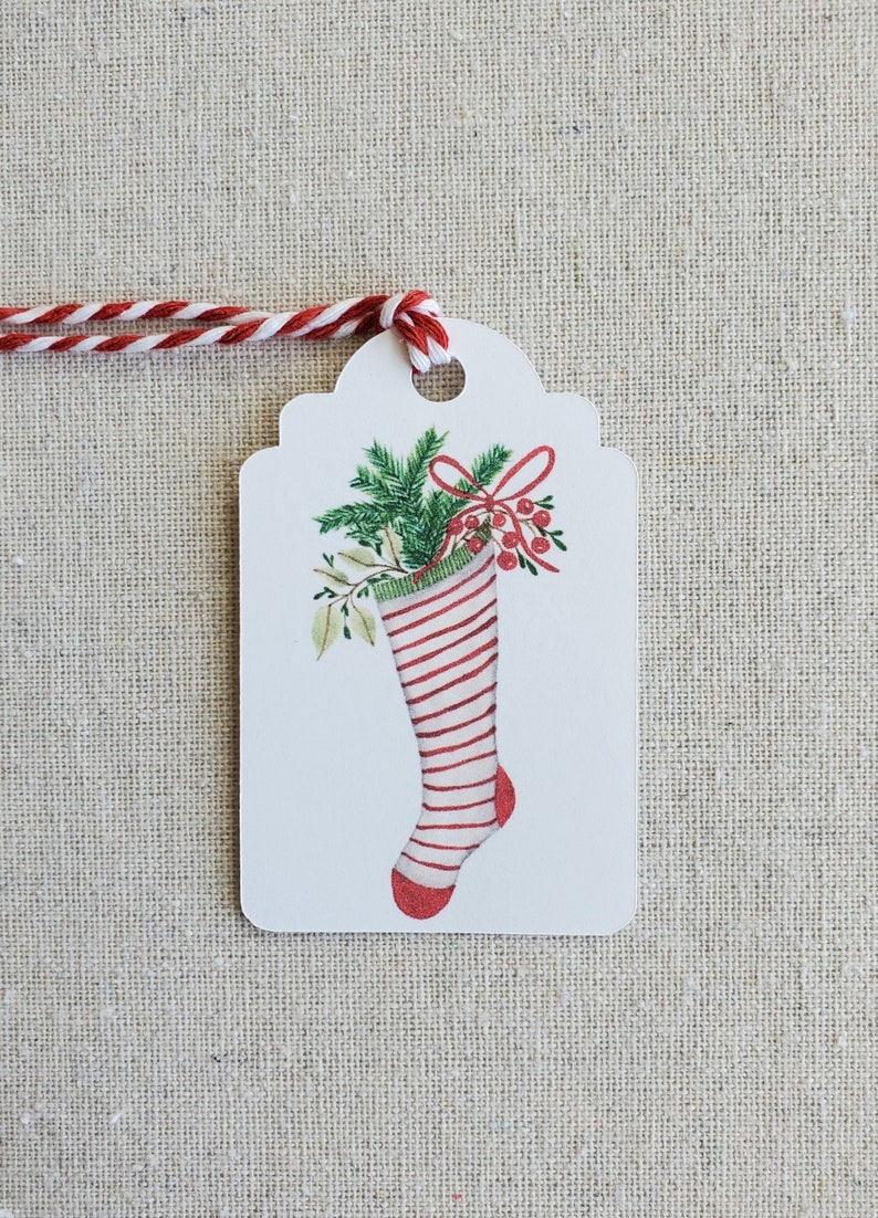 May include: A white gift tag with a red and white striped stocking filled with green holly and red berries. The tag is tied with a red and white twine.
