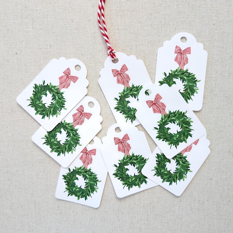 K&ouml;nnte beinhalten: Wei&szlig;e Geschenkanh&auml;nger mit einem gr&uuml;nen Kranz und rotem Schleifendesign. Die Anh&auml;nger sind mit rot-wei&szlig;em Garn verbunden. Eine festliche Weihnachtsdekoration.