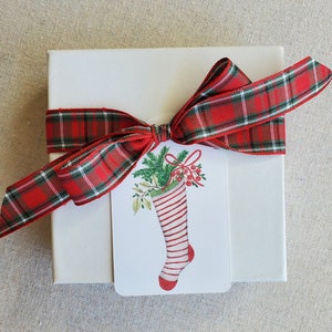 May include: A white gift box with a red and green plaid ribbon tied in a bow. A white gift tag with a red and white striped stocking filled with green holly and red berries is attached to the ribbon.