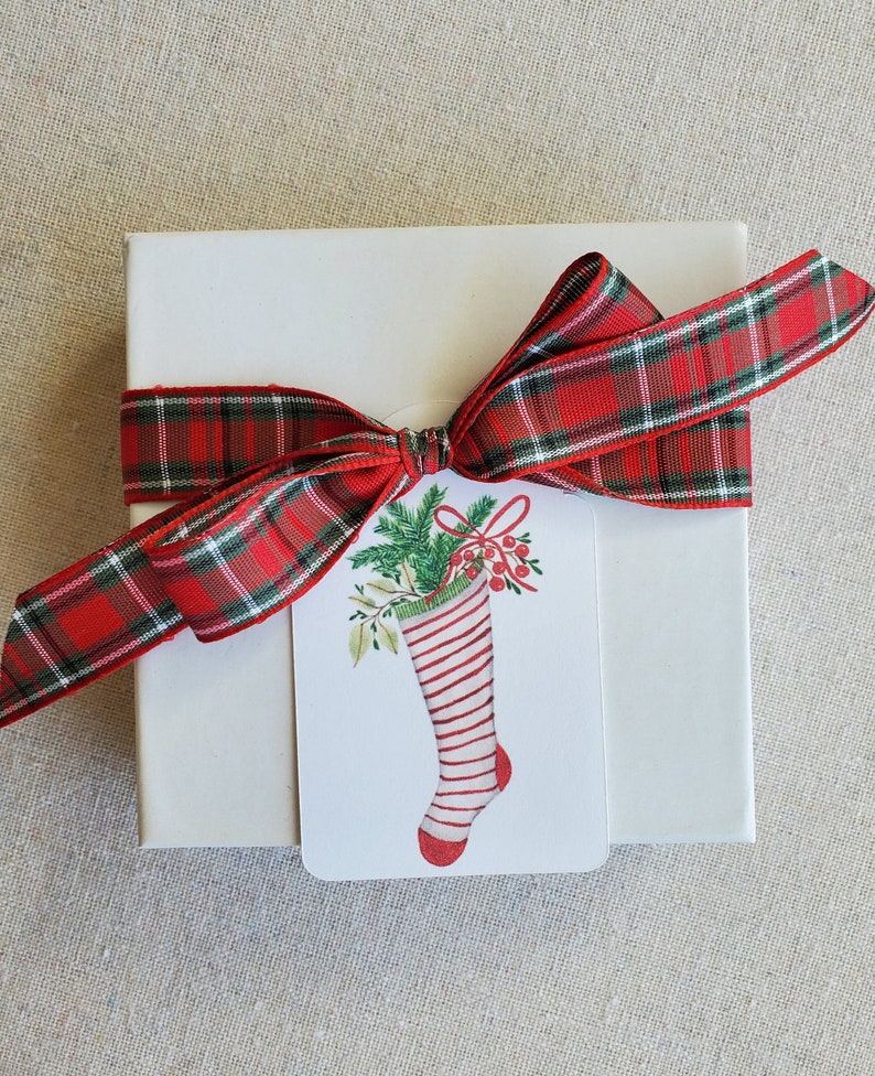 May include: A white gift box with a red and green plaid ribbon tied in a bow. A white gift tag with a red and white striped stocking filled with green holly and red berries is attached to the ribbon.