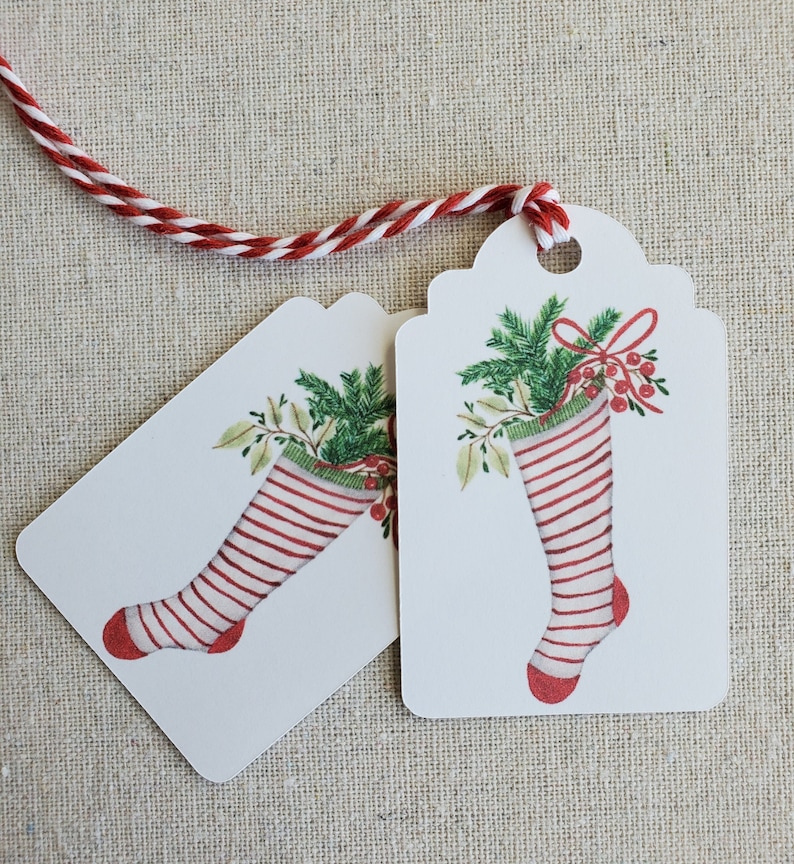 May include: Two white gift tags with red and white striped stockings filled with green holly and red berries. The tags have scalloped edges and a hole punched at the top.