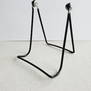 May include: A black metal display stand with a minimalist design. The stand has a curved base and two upright supports with silver-colored, circular accents at the top. The stand is designed to hold a small object.