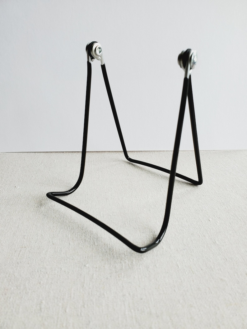 May include: A black metal display stand with a minimalist design. The stand has a curved base and two upright supports with silver-colored, circular accents at the top. The stand is designed to hold a small object.