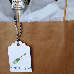 May include: A brown paper gift bag with a white gift tag attached. The tag has a hand-drawn illustration of a champagne bottle popping with confetti and the text "Happy New Year!"
