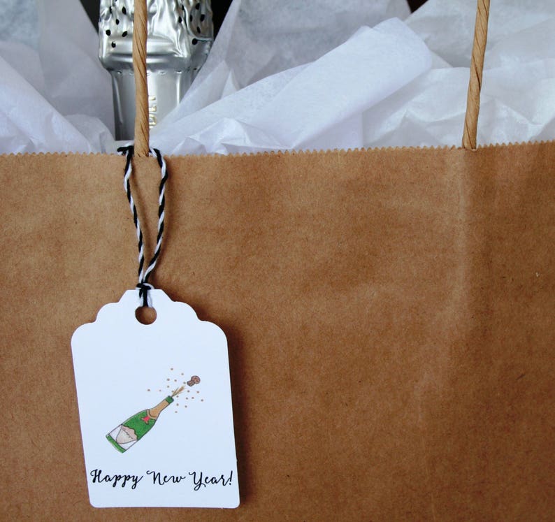 May include: A brown paper gift bag with a white gift tag attached. The tag has a hand-drawn illustration of a champagne bottle popping with confetti and the text "Happy New Year!"