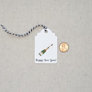 May include: White gift tag with a black and white string. The tag has a drawing of a champagne bottle popping with a cork flying out. The text on the tag reads "Happy New Year!"