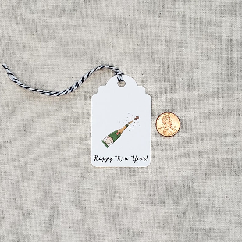 May include: White gift tag with a black and white string. The tag has a drawing of a champagne bottle popping with a cork flying out. The text on the tag reads "Happy New Year!"