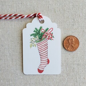 May include: White gift tag with a red and white striped stocking filled with green holly and red berries. The stocking has a green cuff and a red toe. A red bow is tied around the holly.