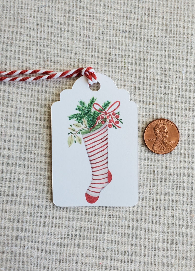 May include: White gift tag with a red and white striped stocking filled with green holly and red berries. The stocking has a green cuff and a red toe. A red bow is tied around the holly.