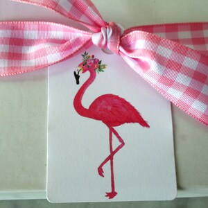 K&ouml;nnte beinhalten: Ein wei&szlig;er Geschenkanh&auml;nger mit einem pinkfarbenen Flamingo, der eine Blumenkrone tr&auml;gt. Der Anh&auml;nger ist mit einem rosa-wei&szlig; karierten Band gebunden.