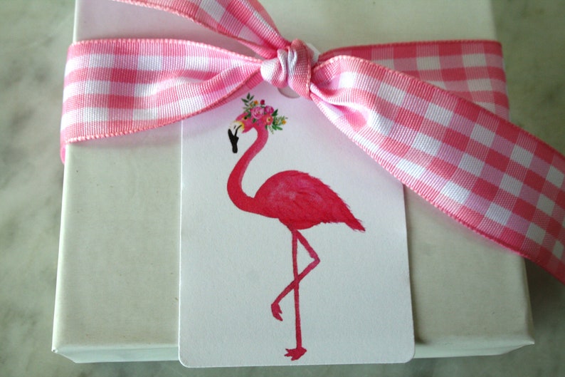 K&ouml;nnte beinhalten: Ein wei&szlig;er Geschenkanh&auml;nger mit einem pinkfarbenen Flamingo, der eine Blumenkrone tr&auml;gt. Der Anh&auml;nger ist mit einem rosa-wei&szlig; karierten Band gebunden.