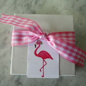 K&ouml;nnte beinhalten: Eine wei&szlig;e Geschenkbox mit einem rosa-wei&szlig;en Gingham-Band, das zu einer Schleife gebunden ist. Ein rosa Flamingo mit einer Blumenkrone befindet sich auf einem Etikett, das an der Schleife befestigt ist.