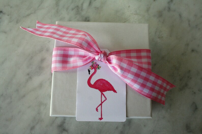 K&ouml;nnte beinhalten: Eine wei&szlig;e Geschenkbox mit einem rosa-wei&szlig;en Gingham-Band, das zu einer Schleife gebunden ist. Ein rosa Flamingo mit einer Blumenkrone befindet sich auf einem Etikett, das an der Schleife befestigt ist.