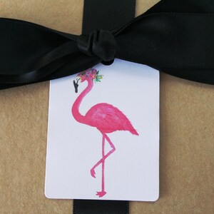 K&ouml;nnte beinhalten: Ein rosa Flamingo mit einer Blumenkrone auf dem Kopf, der auf einem Bein steht. Der Flamingo befindet sich auf einem wei&szlig;en Geschenkanh&auml;nger mit einem schwarzen Band.
