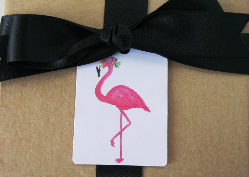 K&ouml;nnte beinhalten: Ein rosa Flamingo mit einer Blumenkrone auf dem Kopf, der auf einem Bein steht. Der Flamingo befindet sich auf einem wei&szlig;en Geschenkanh&auml;nger mit einem schwarzen Band.