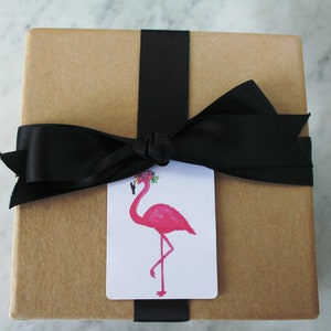 K&ouml;nnte beinhalten: Eine braune Geschenkbox mit einem schwarzen Band, das zu einer Schleife gebunden ist. Ein wei&szlig;es Geschenkanh&auml;nger mit einem rosa Flamingo, der eine Blumenkrone tr&auml;gt, ist am Band befestigt.