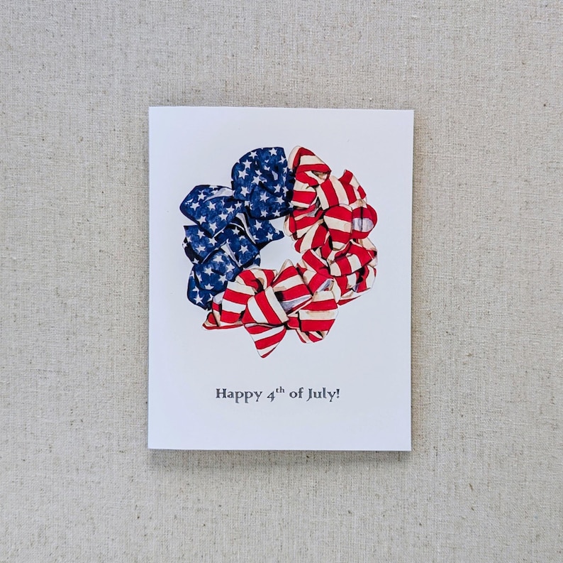 May include: A white card with a red, white, and blue fabric wreath in the shape of a circle. The wreath is made of fabric with the American flag pattern. The text "Happy 4th of July!" is printed below the wreath.