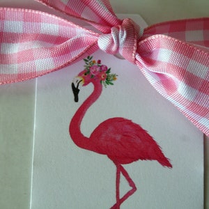 K&ouml;nnte beinhalten: Ein rosa Flamingo mit einer Blumenkrone auf dem Kopf. Der Flamingo steht auf einem wei&szlig;en Hintergrund mit einem rosa-wei&szlig;en Gingham-Band, das um ihn herum gebunden ist.