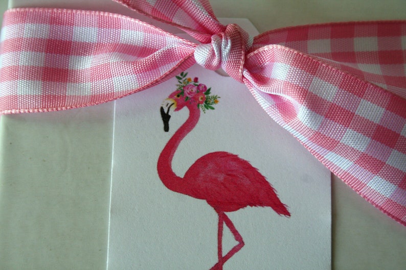 K&ouml;nnte beinhalten: Ein rosa Flamingo mit einer Blumenkrone auf dem Kopf. Der Flamingo steht auf einem wei&szlig;en Hintergrund mit einem rosa-wei&szlig;en Gingham-Band, das um ihn herum gebunden ist.