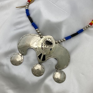 May include: A silver necklace with a large, ornate pendant featuring a stylized design. The pendant has three smaller circles hanging from it. The necklace is made of blue, black, and red beads.