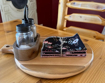 May include: A wooden serving tray with a round, light-colored top and a dark-colored rim. It holds a wooden caddy with glass salt and pepper shakers and a stack of black floral print napkins tied with twine.