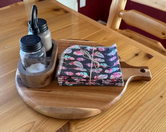 May include: A wooden serving board with a handle holds a stack of feather-patterned napkins tied with twine. A wooden caddy with two glass jars and a metal oil dispenser sits to the left. The jars have black lids.