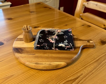 May include: A wooden serving board with a handle holds a stack of black floral print napkins tied with twine. A small wooden container with toothpicks sits to the left. The items are on a wooden table.